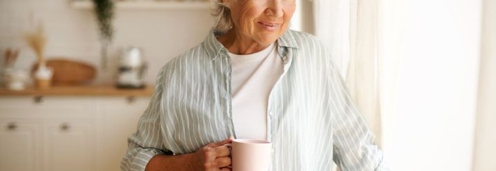 Ältere Person mit einer Tasse blickt lächelnd aus dem Fenster.